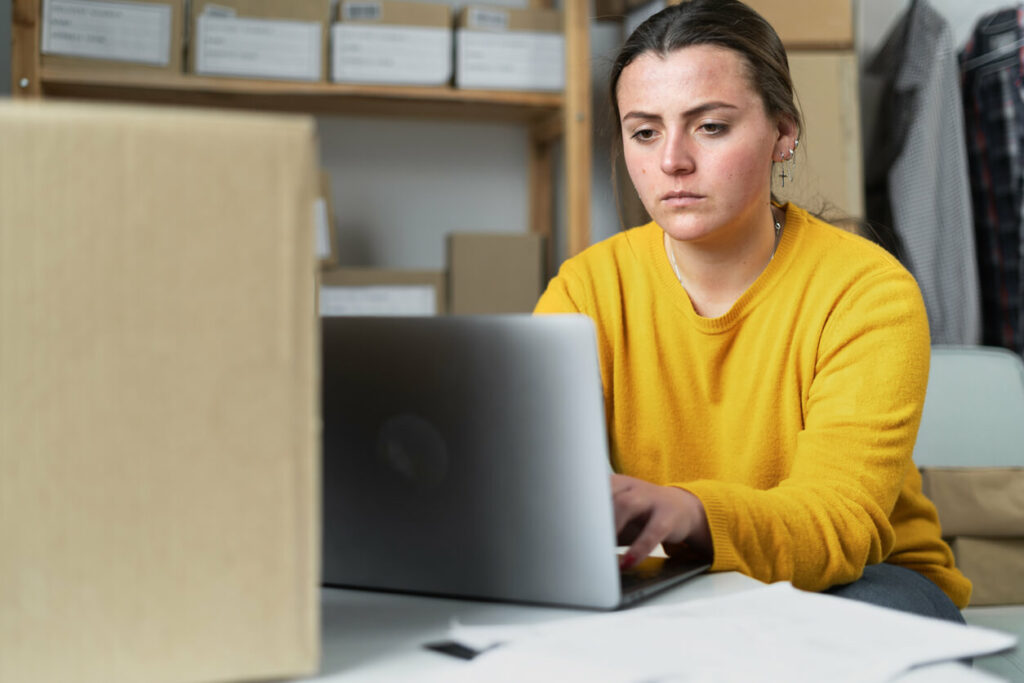 Small business owner working on a laptop with a box in the foreground and boxes on shelves in the background.