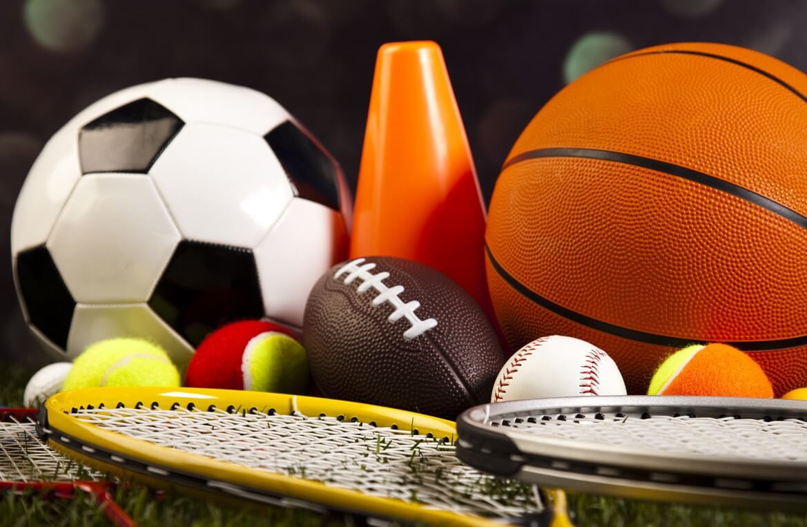 A close-up composition of various sports balls and pieces of sports equipment resting together on grass.